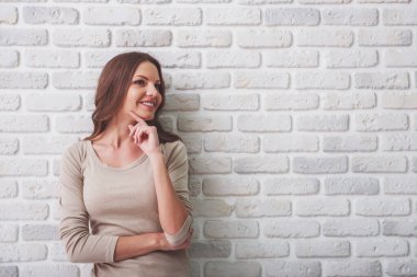 Rahat giysiler içinde güzel genç kadın gülümsüyor ve uzak bakıyor, beyaz tuğla duvara karşı