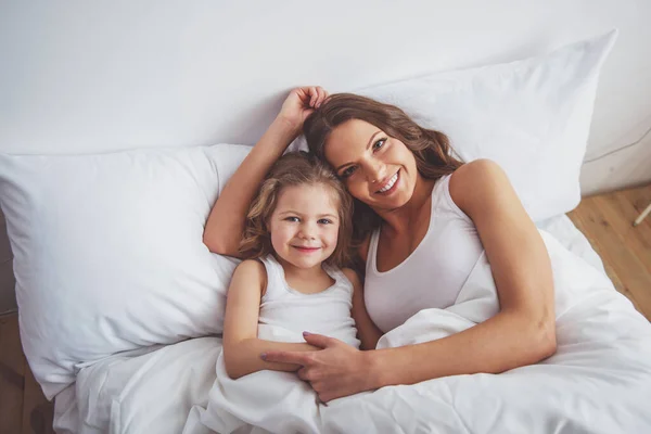 かわいい女の子と彼女の美しい若い母親が寄り添う カメラ目線 ながら自宅のベッドで横になっている笑顔 — ストック写真