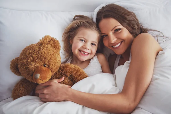 かわいい女の子と彼女の美しい若い母親がカメラ目線し ながら自宅のベッドにテディベアと横になっている笑顔 — ストック写真