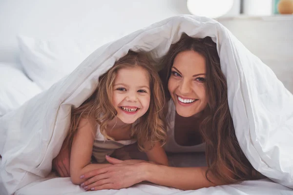 かわいい女の子と彼女の美しい若い母親がカメラ目線し ながら毛布の下のベッドで横になっている笑顔 — ストック写真