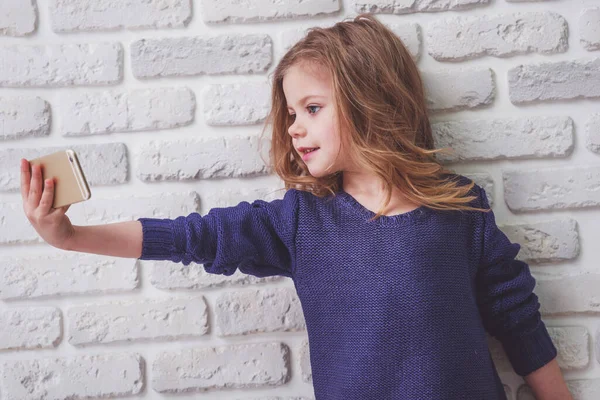 カジュアルな服を着た美しい小さな女の子は 白レンガの壁に対して スマートフォンと笑顔を使用して自分撮りを作っています — ストック写真