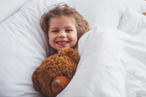 Солодка Дівчинка Обіймає Плюшевого Ведмедя Дивлячись Камеру Посміхаючись Лежачи Ліжку — стокове фото