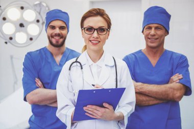 Güzel kadın doktor ön planda ve iki erkek kameraya bakıyor ve gülümsüyor, modern bir ameliyat odasında duran