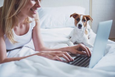 Rahat kıyafetler kız güzel bir laptop kullanıyor ve onun sevimli köpek yatakta yatan süre gülümseyen, yakın çekim. Evde beslenen hayvan kameraya bakıyor