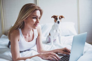 Güzel sarışın kız rahat kıyafetler içinde bir dizüstü bilgisayar kullanarak ve onun sevimli köpek yatakta yatan süre gülümseyen. Evde beslenen hayvan kameraya bakıyor