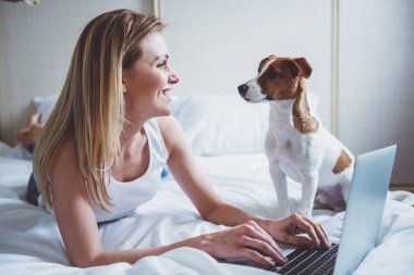 Güzel kızı rahat kıyafetler gülümseyen ve onun sevimli köpek yatakta yatıyordu süre seyir, bir dizüstü bilgisayar kullanarak