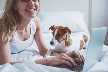 Güzel sarışın kız rahat kıyafetler içinde kameraya bakarak, bir dizüstü bilgisayar kullanarak ve onun sevimli köpek yatakta yatan süre gülümseyen, yakın çekim