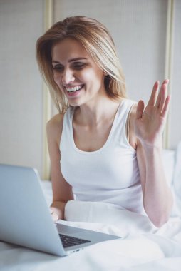 Güzel sarışın kız sallayarak, bir dizüstü bilgisayar kullanarak ve yatakta otururken gülümseyen