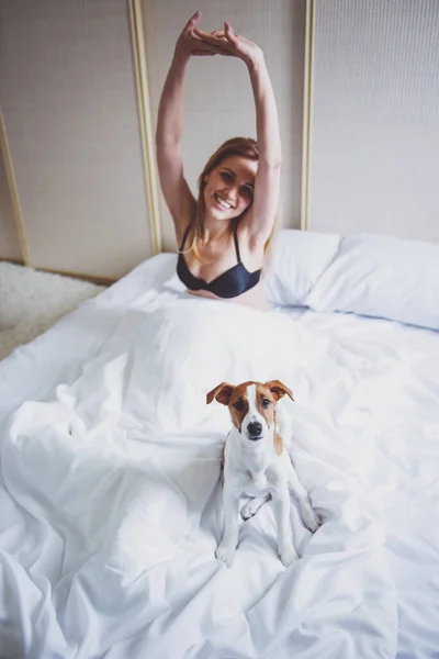 黒下着で美しいブロンドの女の子はストレッチと ベッドの上に座って笑っています 彼女のかわいい犬はカメラを目線します — ストック写真