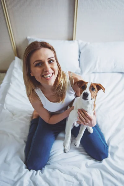私服かわいい愛犬を抱いて カメラを見て ベッドの上に座って笑顔で美しいブロンドの女の子のトップ ビュー — ストック写真