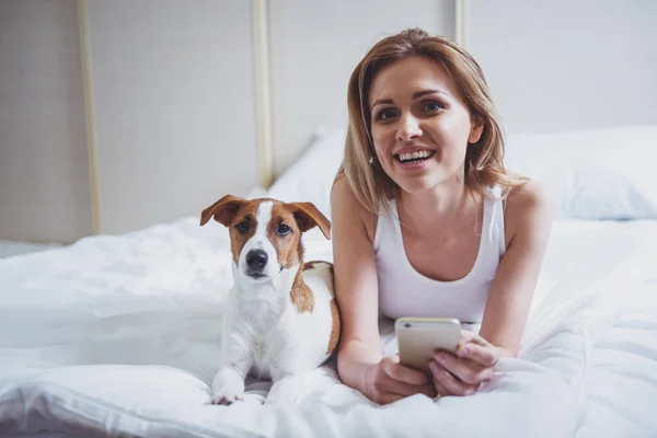 カジュアルな服で美しいブロンドの女の子はスマート フォンを使用と彼女のかわいい犬の両方のカメラ目線のベッドの上に横たわっている間笑顔 — ストック写真