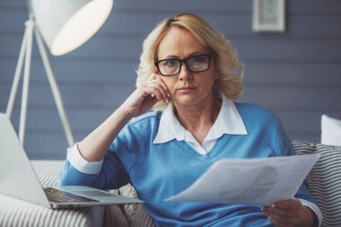 Rahat kıyafetler ve gözlük güzel yaşlı kadın evde laptop ile çalışırken kağıtları tutuyor