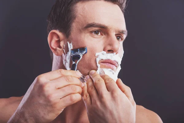 英俊的裸体男人剃须使用一把剃刀 看向别处 在深色背景上的肖像 — 图库照片