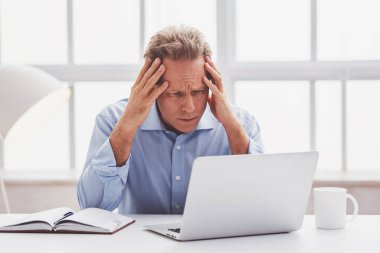 Yakışıklı yorgun orta yaşlı işadamı klasik gömlek bir dizüstü bilgisayar kullanmak ve ofiste çalışırken kafasına ellerini tutmak