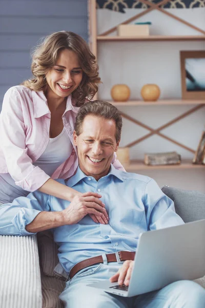 快乐美丽的夫妇正在使用一台笔记本电脑 拥抱和微笑着坐在家里的沙发上 — 图库照片