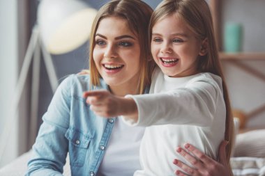 Güzel genç anne ve küçük kızı sarılma, gülümseyen, seyir ve uzak evde kanepede otururken işaret