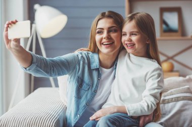 Güzel genç anne ve küçük kızı bir akıllı telefon kullanarak ve evde kanepede otururken gülümseyen bir selfie yapıyoruz