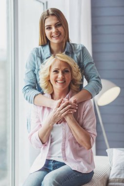 Güzel olgun anne ve kızını sarılma, kameraya bakıyor ve gülümsüyor, evde koltukta oturan portre