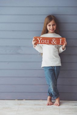 Ahşap bir plaka tutan, kameraya bakan ve gülümseyen, gri arka plana karşı duran güzel küçük kızın tam uzunlukta portre