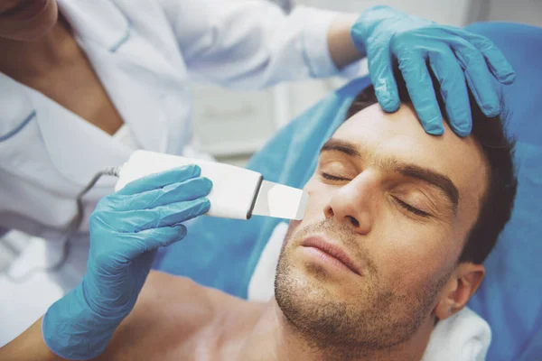 英俊的男人正在脸上的皮肤治疗 医用手套的医生正在采用一种现代设备的程序 — 图库照片