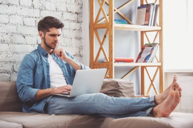 Çekici dalgın genç adam evde kanepede otururken bir laptop kullanıyor