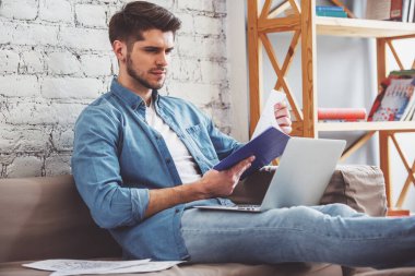 Çekici bir genç adam çalışma, belgelerin incelenmesi ve evde kanepede otururken bir dizüstü bilgisayar kullanarak