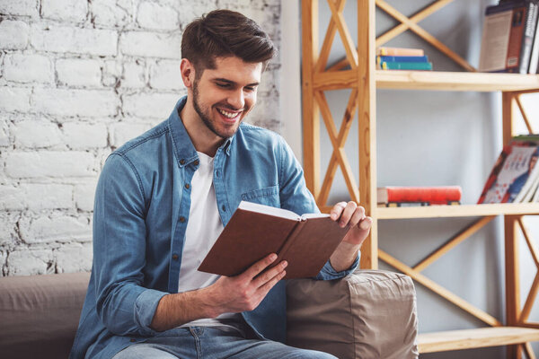 Attractive young man is reading a book and smiling while sitting on sofa at home