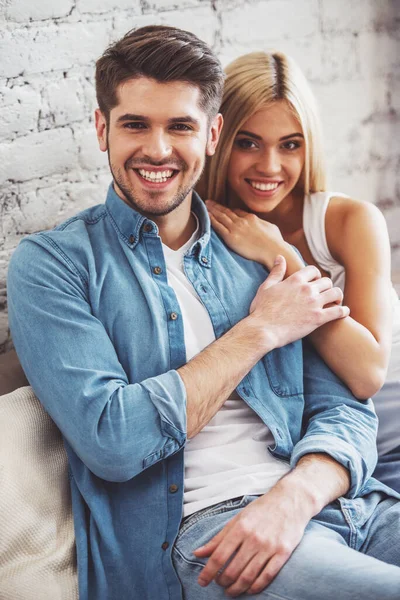 美しい若いカップルがハグ カメラ目線と自宅のソファに腰掛けながら笑顔 — ストック写真