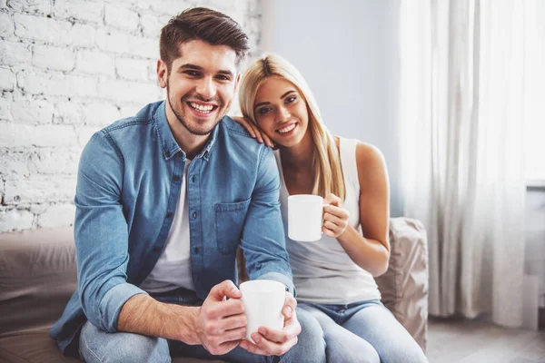 stock image Beautiful young couple is holding cups of drink, looking at camera and smiling while sitting on sofa at home