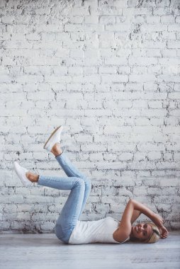 Rahat giysiler içinde Çekici kız kamera bakıyor ve gülümseyerek, beyaz tuğla duvara karşı katta yukarı bacaklar ile yalan