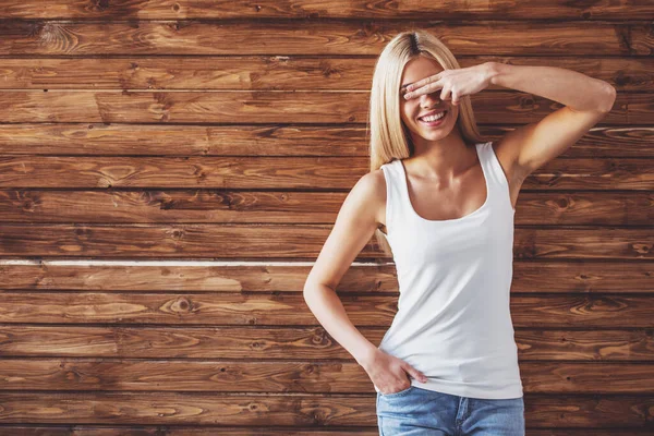 カジュアルな服を着た魅力的な若い女性は 木製の壁に立って 彼女の目を覆い 笑顔です — ストック写真
