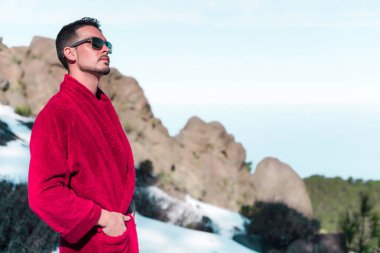 Kırmızı uzun bornozlu genç adam Mauntain tepesinde karlı güneşli günün tadını çıkarıyor.