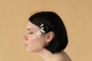 Profilde genç ve güzel bir kadının portresi, kulağının arkasında beyaz bir çiçek, sarı bir duvarın yanında.