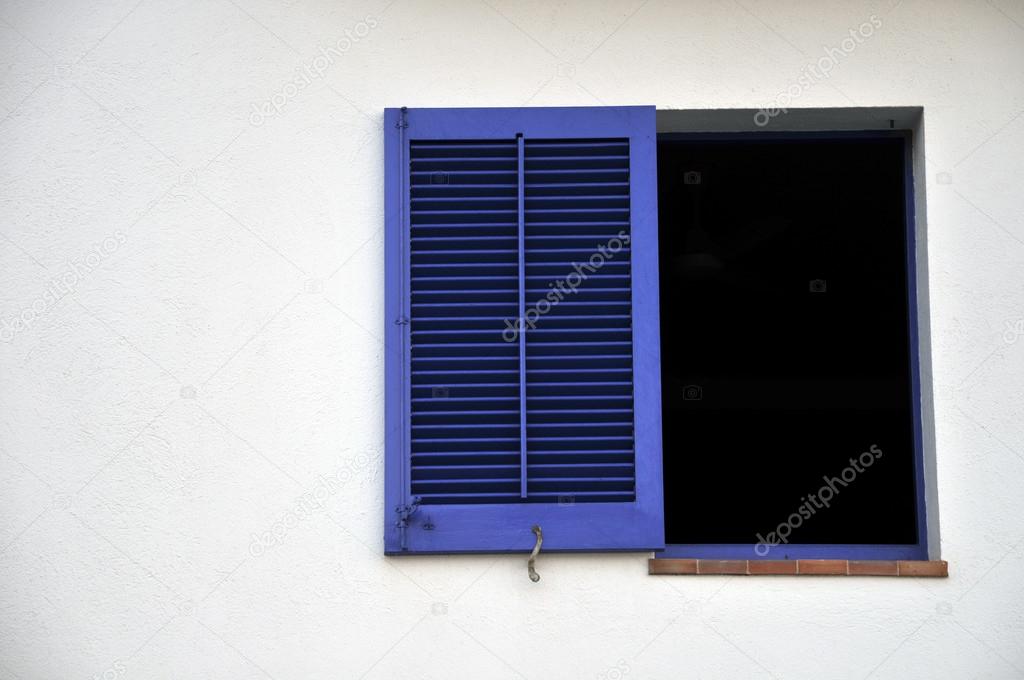 A blue window shutter on a white wall. Black window frame ideal to place your custom art for southern mediterranean travel concepts