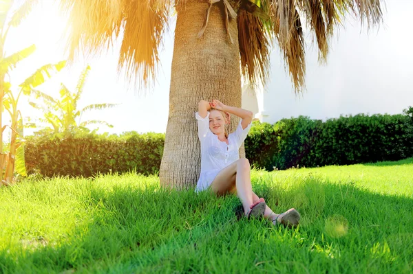 Meisje, zittend op het gras onder een palmboom — Stockfoto