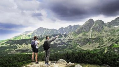 İki genç turist bir erkek ve bir kadın yazın vahşi doğada dinlenirken kayaların üzerinde manzaralı bir dağ manzarası seyrediyorlar.