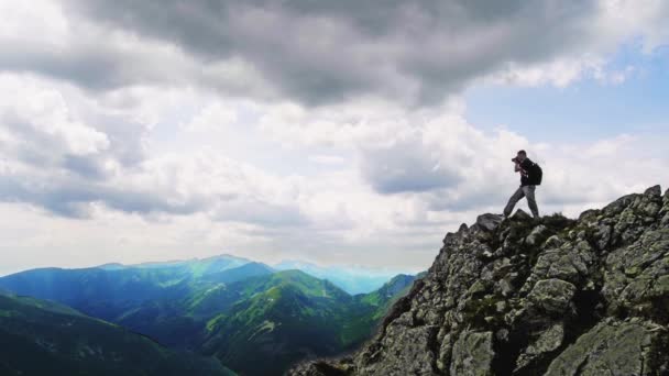 Красивый молодой человек гуляет по горам, исследует природу, стоит на скале и фотографируется с камерой, фотографирует окружающую его природу.. — стоковое видео