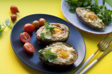Yumurta, peynir, mısır salatası ve domatesli fırında avokado, sarı arka planda siyah ve beyaz tabaklar. Avokadoyla kahvaltı. Altın bıçak ve çatal.. 