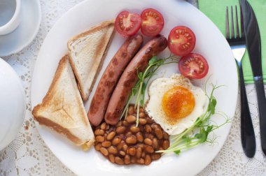 Yağda yumurta, sosis, tost, domates, bezelye filizi ve beyaz tabakta fasulye ile İngiliz kahvaltısı. Geleneksel İngiliz kahvaltısı çay ve çaydanlıkla beyaz dantel masa örtüsü üzerinde çatal ve bıçakla. Üst görünüm