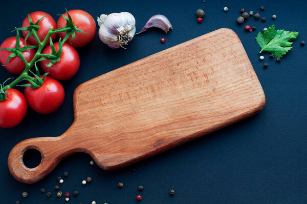 Empty wooden board with food ingredients on black background. Garlic, tomatoes, spices, pepper, parsley around wooden board