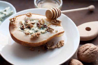 Dorblu peynirli armut, bal ve fındıklı beyaz tabakta koyu kahverengi arka planda. Tahta tahtada armut ve tahta bal çubuğu. Kırsal biçim