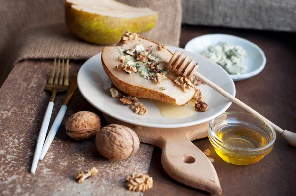 Koyu kahverengi ve ahşap arka planda beyaz bir tabakta Dorblu peyniri, balı ve cevizli armut. Ahşap tahta tahtada armut, tahta bal sopası, bıçak ve çatal. Kırsal biçim