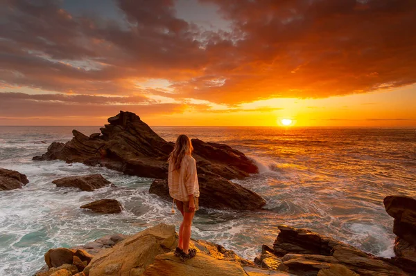 カジュアルなクリームリネンのシャツとショートパンツを着た女性は 波が彼女の周りにクラッシュとして海の上に鮮やかな赤い日の出を見て岩の上に立っています ロイヤリティフリーのストック写真