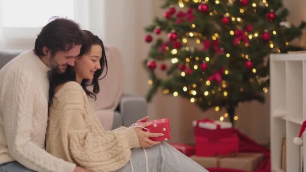 Пара святкує Новий рік біля новорічної ялинки вдома, дарує подарунки в подарункових коробках — стокове відео