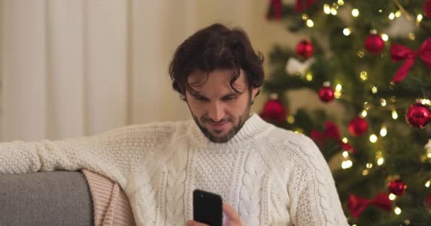Человек празднует Новый год возле елки дома, используя смартфон — стоковое видео
