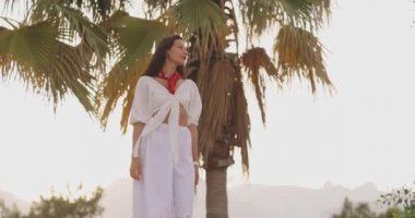 Yaz tatili boyunca arka planda palmiye ağaçlarına karşı şık plaj kıyafeti giyen kadın.