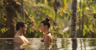 Panoramic photo of young adult couple in love swimming together in open-air infinity pool enjoying tropical vacation on villa looking to each other against rainforest on sunset. High quality 4k