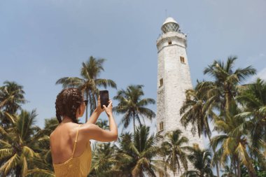 Romantic woman traveler in dress taking photo on smartphone of famous landmarks of Sri Lanka country. Young girl standing and looking on Dondra lighthouse. High quality travel photo