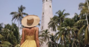 Back view of romantic woman traveler in dress and straw hat visiting famous landmarks of Sri Lanka country standing and looking on Dondra lighthouse. High quality 4k slow motion footage
