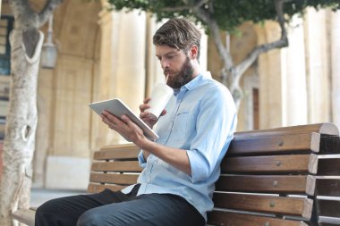 Bankta oturan genç adam bir tabletten okuyor.